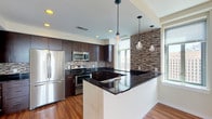 Located in the heart of St. Louis, this two-floor residence at 1212-28 Olive St, Unit 2004, offers a well-appointed urban living experience with contemporary finishes throughout. The open-concept kitchen is a standout feature, showcasing rich dark cabinetry, sleek black granite countertops, a mosaic tile backsplash, and a full suite of stainless steel appliances. A central island with pendant lighting serves as both a functional workspace and a visual anchor, while large windows frame striking downtown St. Louis views. Warm hardwood flooring extends through the main living and dining areas, filling the space with natural light and a polished, modern feel. A sculptural multi-bulb chandelier adds an artistic touch to the dining area. The living area on the lower level features plush carpeting, wall sconces, and a welcoming, versatile layout. The primary bathroom is finished with large-format travertine tile, a soaking tub, a glass-enclosed shower, and generous floor space. A second bathroom mirrors this refined aesthetic with dark wood cabinetry and travertine tile surround. Upstairs, carpeted bedrooms offer walk-in closet access and open onto the stairwell landing. Throughout both levels, the design balances contemporary urban style with comfortable, functional living.