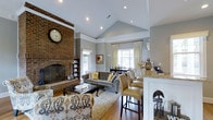 This single-story home showcases a thoughtfully designed open-concept layout that connects the living, dining, and kitchen areas into one cohesive, flowing space. The living room is anchored by a striking floor-to-ceiling brick fireplace, complete with a decorative hearth, adding a classic architectural focal point to the room. Soaring vaulted ceilings with recessed lighting enhance the sense of openness, while soft gray walls create a neutral, welcoming palette throughout.

Warm hardwood flooring runs continuously across the space, contributing to its unified and airy feel. The living area is furnished with a tufted gray sofa, patterned accent chairs, and a traditional coffee table atop a light area rug. Large windows dressed with sheer curtains allow generous natural light to fill the room.

The dining area sits adjacent, accommodating a dark wood table with seating, positioned near additional windows. The kitchen flows naturally from the main living space, featuring white cabinetry, light granite countertops, and a raised breakfast bar with barstool seating — making it ideal for both casual dining and entertaining.