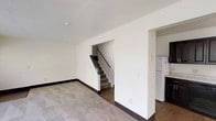 This two-floor space offers a clean, straightforward layout designed for practical everyday living. The main level features a spacious carpeted living area with neutral walls and dark baseboards that provide a subtle contrast throughout. A textured ceiling adds a traditional architectural touch overhead. An open staircase with a dark wood handrail leads to the upper floor, conveniently positioned within the main living space for easy access between levels. Adjacent to the living area, the kitchen is separated by a partial wall, offering a degree of definition while maintaining an open feel. The kitchen is equipped with a white refrigerator and a dark appliance, set against hard flooring that contrasts with the carpeted living zone. The overall color palette is neutral and understated, with white walls and light tones creating a bright, airy atmosphere throughout. The layout maximizes usable space across both floors, making it well-suited for a variety of configurations. The clean lines and simple finishes give the space a versatile foundation that can be easily adapted to suit different needs and styles.