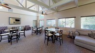 This single-floor community space is defined by its striking architectural centerpiece: a soaring vaulted great room with exposed white-painted timber trusses and ceiling fans that draw the eye upward, creating an airy, open atmosphere. Large windows along the perimeter flood the interior with natural light and offer views of the surrounding greenery. The main gathering area accommodates multiple dining tables and chairs arranged beneath the dramatic roofline, making it well-suited for group events and community gatherings. Adjacent to the dining area is a comfortable lounge space featuring plush upholstered armchairs, patterned carpeting, and a ceiling fan — a relaxed setting ideal for casual conversation. A separate sitting area provides a cozy retreat with a large sectional sofa, wall-mounted television, and a tiled fireplace as its focal point. The space includes two kitchens, each outfitted with rich cherry wood cabinetry, granite-style countertops, black appliances including a range, microwave, dishwasher, and refrigerator, offering ample preparation space for catered events or communal use. Together, these areas combine practical amenities with warm, welcoming design throughout.
