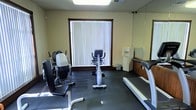This single-floor fitness room provides a functional and well-equipped workout space designed to support a range of exercise routines. The room features durable rubber flooring throughout, offering both safety and stability for physical activity. A large window dressed with vertical blinds allows natural light to filter into the space, creating a bright and comfortable atmosphere.

The room is outfitted with a selection of cardio and strength training equipment, including a stationary upright exercise bike, a treadmill, and an adjustable resistance training machine, making it suitable for a variety of fitness regimens. A water cooler station is conveniently positioned along one wall, ensuring easy access to hydration during workouts. A full-length mirror runs along one wall, allowing users to monitor their form and technique.

The space is finished with neutral, light-toned walls and features recessed overhead lighting to supplement the natural light. A compact counter area adjacent to the mirror provides a small utility surface. The overall layout is efficiently organized to maximize the usable floor space while keeping equipment accessible. This fitness room offers a straightforward, no-frills environment focused on delivering a practical and comfortable exercise experience.