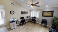 This single-floor office space is thoughtfully designed to support a functional and professional work environment. The room features an L-shaped built-in granite-top workstation that spans two walls, providing ample desk space for multiple workstations — each equipped with a computer monitor and chair. A multifunction printer/copier station is conveniently positioned along one wall, while an additional guest chair offers seating for visitors. The flooring consists of neutral-toned stone tile, adding a polished, durable foundation throughout the space. Crown molding runs along the upper perimeter of the room, lending a refined architectural detail that elevates the overall aesthetic. A ceiling fan with an integrated light fixture is centrally mounted, ensuring consistent air circulation and ambient lighting. Recessed ceiling lights provide additional illumination, complementing the natural light that filters through a plantation-shuttered window, which allows for adjustable light control and privacy. Warm, neutral wall tones create a calm and focused atmosphere, accented by wall-mounted artwork. The overall design balances practicality with a polished, professional ambiance well-suited for office or administrative use.