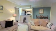 This single-floor residence offers an open-concept layout that thoughtfully connects the main living areas into one cohesive space. The living room serves as the heart of the home, featuring plush carpeted flooring and soft, neutral-toned walls that create a warm and welcoming atmosphere. A peninsula-style kitchen sits at the center of the layout, featuring light wood cabinetry, a full suite of appliances including a refrigerator and electric range, and a breakfast bar with barstool seating — ideal for casual dining or entertaining. Recessed lighting and under-cabinet illumination keep the kitchen bright and functional. An accent wall in a muted teal hue adds a subtle pop of color to the dining area, which accommodates a table and chairs for everyday meals. The transition between spaces is smooth and intuitive, with an open sightline from the living room through the kitchen and into the dining area. A doorway off the living room provides access to additional private areas of the residence. The overall ambiance is cozy yet practical, with a clean, contemporary aesthetic accented by soft lighting and tasteful decor throughout.