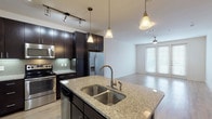This well-appointed single-floor residence at Agave Pass in San Antonio, Texas, offers a thoughtfully designed open-concept layout that seamlessly connects the kitchen, dining, and living areas. The gourmet kitchen is a standout feature, showcasing rich dark espresso cabinetry that extends to the ceiling for ample storage, complemented by sleek granite countertops with subtle veining. A generously sized central island with a double undermount stainless steel sink serves as a practical workspace and natural gathering point. Stainless steel appliances, including a French door refrigerator, electric range, and built-in microwave, complete the kitchen's modern aesthetic. A tasteful tile backsplash and track lighting add refined detail overhead, while elegant pendant lights illuminate the island. The open living area flows naturally from the kitchen, featuring light wood-style flooring, a ceiling fan, and floor-to-ceiling French doors that invite natural light and provide outdoor access. The bedroom is carpeted for comfort and is accompanied by a bathroom with tile flooring and a spacious walk-in closet with dual hanging rods. Throughout, the neutral color palette and clean lines create a bright, contemporary ambiance suited for modern living.