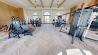 This expansive 2,350 square foot fitness center is a testament to modern design and functionality. The space boasts a single-floor layout, maximizing accessibility and flow. Architectural highlights include a striking coffered ceiling with exposed wooden beams, adding warmth and character to the pristine white interior.

Large arched windows dominate the walls, flooding the space with natural light and offering picturesque views of the surrounding landscape. These windows not only enhance the aesthetic appeal but also create an open, airy atmosphere conducive to invigorating workouts.

The center is equipped with a comprehensive array of state-of-the-art fitness machines, including cardio equipment, weight training stations, and specialized workout areas. The layout is thoughtfully organized into distinct zones, catering to various exercise preferences and intensities.

The herringbone pattern of the light wood flooring adds a touch of sophistication while providing a stable surface for workouts. Strategically placed recessed lighting ensures even illumination throughout the space.

A notable feature is the large decorative wall art, which adds a vibrant splash of color and inspiration to the otherwise neutral palette. This careful balance of form and function creates an inviting environment that encourages health and wellness pursuits in a luxurious setting.