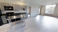This Type C residence features 2 bedrooms and 2 bathrooms across a single thoughtfully arranged floor. The open-concept layout seamlessly connects the kitchen and living areas, creating a bright and functional space that feels expansive and welcoming. The kitchen is outfitted with contemporary gray cabinetry, dark granite countertops, and a stainless steel sink, complemented by black appliances. A movable stainless steel utility cart adds a versatile, modern touch to the culinary space. Light-toned luxury vinyl plank flooring extends throughout the main living area, lending a clean, cohesive aesthetic that reflects natural light beautifully. The living area is generously proportioned, with neutral wall tones and multiple windows that invite natural light and provide views to the outdoors. Recessed ceiling vents ensure efficient climate control throughout. Doorways lead to the private bedroom and bathroom quarters, maintaining a clear separation between living and sleeping spaces. The overall ambiance is modern and move-in ready, with a neutral palette that offers flexibility for personalization. This residence is an ideal choice for those seeking a practical yet stylish home with a well-balanced floor plan.