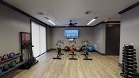 The Gentry's fitness center is a thoughtfully designed, single-floor space that accommodates a wide range of workout styles and training needs. The room is finished with light wood-tone luxury vinyl plank flooring throughout, set against warm gray walls with dark trim accents that give the space a modern, polished aesthetic. Overhead, linear LED lighting and ceiling fans ensure the environment remains well-lit and comfortable during workouts.

The space is divided into distinct functional zones. A dedicated cycling area houses multiple spin bikes arranged facing a wall-mounted flat-screen television, ideal for guided or self-directed cardio sessions. Stability balls of various sizes are stored nearby for core and balance training. A large roller shade-equipped window wall brings in natural light while maintaining privacy.

The strength and functional training zone features a multi-use cable machine, a full dumbbell rack with a comprehensive weight range, kettlebells of varying sizes, resistance bands, body bars, and exercise mats—all neatly organized on dedicated racks. A full-length mirror along one wall enhances spatial awareness during workouts. Punching bags add a boxing and functional fitness element to the space.

An adjacent hallway area provides additional storage hooks and access to private rooms, completing a well-rounded and professionally appointed fitness facility.