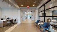 This thoughtfully designed single-floor amenity space combines refined aesthetics with functional versatility. The entry corridor sets the tone immediately, featuring a striking herringbone-patterned light wood floor, a textured decorative ceiling with recessed lighting, and a glass-framed wall that frames views into adjacent areas. A staffed reception desk with modern task lighting sits just off the main corridor, offering a welcoming point of contact upon arrival.

The central lounge is a standout feature, boasting soaring double-height ceilings lined with floor-to-ceiling black-framed windows that flood the space with natural light and provide views of the outdoor surroundings. Elegantly curated furnishings — including plush sofas, leather seating, and gold-accented side tables — are arranged atop layered area rugs, anchoring the space with warmth and sophistication. Bold original artwork adorns the walls, adding an artistic dimension throughout.

A dedicated lounge nook showcases an illuminated accent wall mural and gallery-style framed artwork, creating an intimate retreat within the larger space. The full kitchen area features dark cabinetry with open shelving, stainless steel appliances, a marble-topped island with bar seating, and pendant lighting with brushed gold fixtures — ideal for social gatherings. Barn-style sliding doors and clean white walls complete the polished, contemporary character of this amenity space.