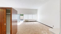 This efficient single-floor unit is thoughtfully laid out to maximize every square foot of its compact footprint. The entrance leads into a bright, open-plan living and dining area that serves as the heart of the space. Large floor-to-ceiling windows dressed with vertical blinds span the length of the main room, flooding the carpeted interior with abundant natural light and creating a spacious, airy atmosphere. A sliding glass door connects the living area directly to the private outdoor patio, which is enclosed by a distinctive decorative breeze-block wall and wood fencing, offering a secluded retreat with views of mature trees above.

The kitchen is neatly framed within an alcove and features white cabinetry, a light-toned countertop with a breakfast bar overhang, a full-size refrigerator, a sink, and a stovetop — all finished in a clean, utilitarian style. A closet with a dark wood wardrobe unit provides practical storage adjacent to the living area.

The bathroom is finished in classic white, with small mosaic floor tiles, white square wall tiles surrounding the shower-over-tub combination, and a simple vanity with a dark countertop. The overall aesthetic throughout is clean and minimal, with white walls, neutral carpeting, and tile flooring in the kitchen and bath contributing to a bright, understated character.