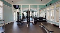 Located at 101 Breakers Dr in Myrtle Beach, South Carolina, this fitness center offers a well-equipped, single-floor exercise facility designed to support a comprehensive workout experience. The interior is characterized by a soothing aqua-toned color palette, crisp white crown molding, and vaulted ceilings that lend an open, airy feel to the space. Recessed lighting throughout ensures the room is well-lit, while multiple large windows with plantation shutters invite ample natural light and views of the surrounding greenery.

The facility is outfitted with a diverse range of cardio machines, including treadmills, an elliptical, and a stationary bike, providing options for varied aerobic training. Strength training is well-supported with a multi-station cable machine, weight benches, and a fully stocked dumbbell rack. A dedicated wall-mounted ball rack holds an assortment of medicine and stability balls, catering to functional fitness routines. Wall-mounted flat-screen televisions are positioned throughout the space for entertainment during workouts.

Durable rubber flooring covers the entire workout area, offering both safety and comfort. Full-length mirrors along one wall enhance the sense of space while allowing users to monitor their form. A water dispenser is conveniently available on-site. The overall ambiance is clean, organized, and welcoming, making this fitness center a practical and pleasant environment for exercise.