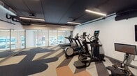 This contemporary fitness center is thoughtfully designed to accommodate a wide range of workout styles across a single, well-organized floor. The space is divided into distinct zones, each serving a specific training purpose. The cardio area is equipped with multiple treadmills, elliptical machines, and stationary bikes, including an Aviron rowing machine, all arranged to maximize usable floor space. A functional training zone features a punching bag, TRX suspension straps, and plyo boxes, providing ample room for high-intensity and bodyweight workouts.

The interior design strikes a bold visual tone, with eye-catching geometric and abstract patterned flooring in earthy tones of orange, tan, grey, and deep teal. The ceiling is finished in a dark, matte black with exposed ductwork and industrial-style linear LED lighting that provides bright, even illumination throughout. White walls keep the space feeling open and clean, while large floor-to-ceiling glass windows along one wall flood the cardio zone with natural light and offer views to the street outside. Full-length mirrors are incorporated near the glass panels, enhancing the sense of depth and space. The overall ambiance is energetic and modern, creating a motivating environment well-suited for both casual users and dedicated fitness enthusiasts.