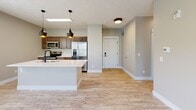 This single-floor space showcases a clean, modern design with a neutral warm-beige and gray color palette carried consistently throughout. Light wood-style plank flooring extends across the main living areas and bedrooms, adding warmth and continuity to the open layout. The kitchen is a functional centerpiece, featuring rich medium-toned wood cabinetry, a full suite of stainless steel appliances including a refrigerator with ice maker, electric range, and built-in microwave. A spacious island with a white countertop and double basin sink anchors the kitchen, illuminated by a pair of stylish black pendant lights with gold interiors. The kitchen flows seamlessly into the open living area, which benefits from multiple windows providing ample natural light and views of surrounding greenery. A front entry door with a modern smart lock leads directly into this open living space. The bedroom areas offer generous proportions with light-filled windows and the same durable flooring throughout. The bathroom includes a full tub and shower combination with white surround, complemented by matte black fixtures that echo the design accents found throughout the unit. The overall ambiance is bright, clean, and move-in ready.
