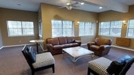This single-story lounge space offers a comfortable and well-appointed environment designed for casual gatherings and relaxation. The room is anchored by a rich brown leather sofa and matching armchair, complemented by a pair of stylish houndstooth-patterned ottomans that add a classic design contrast. A sleek, mirrored coffee table with an ornate metal frame serves as the centerpiece of the seating arrangement, enhancing the room's refined character.

The warm, golden-tan walls create an inviting ambiance, while the carpeted flooring adds comfort underfoot. A ceiling fan with a light fixture is centrally positioned overhead, contributing both to the room's ventilation and its well-lit atmosphere. Recessed lighting along the ceiling provides additional illumination throughout the space.

Multiple large, grid-paned windows span the walls, flooding the room with generous natural light and offering views of lush greenery outside. A wall-mounted sconce adds a subtle decorative accent. A small bistro-style side table near the window provides additional functional surface space. Overall, the room presents a welcoming, traditionally styled lounge ideal for social interaction.