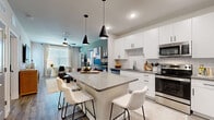 This single-floor, 2-bedroom, 2-bathroom residence offers a cohesive modern design throughout its efficiently utilized layout. The open-concept kitchen is a standout feature, showcasing white shaker cabinetry, a herringbone-patterned tile backsplash, dark countertops, stainless steel appliances, and a central island with seating for four beneath striking black pendant lights — flowing seamlessly into the living and dining areas. The living room is bright and inviting, with large windows, wood-look flooring, and a ceiling fan overhead. The primary bedroom is a comfortable retreat with plush carpeting, a ceiling fan, large windows with drapes, and access to a dedicated closet. The en-suite primary bathroom features a double-sink vanity with white cabinetry, gray countertops, and large mirrors. The second bathroom offers a full tub-shower combo with large-format tile surround and matching white vanity. A second bedroom provides additional flexibility. Two closets offer practical storage throughout. The overall aesthetic is clean and contemporary, with a neutral palette, wood-look flooring in main areas, and consistent modern hardware and fixtures tying the entire space together.