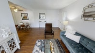 This single-floor space presents an open and well-proportioned layout that seamlessly connects the living and dining areas. Rich, dark hardwood flooring runs throughout, providing a warm and grounding foundation that contrasts elegantly with the bright white walls. The living area is anchored by a tufted blue sofa, a glass-top coffee table with natural wood accents, and a distinctive dark wood reclining chair, creating an inviting and eclectic seating arrangement. A patterned area rug defines the space and adds textural interest underfoot. Wall-mounted framed artwork contributes a curated, finished look to the room. Adjacent to the living area, the dining space accommodates a round table with mid-century modern style chairs, making it ideal for casual meals. A decorative console table positioned along the wall adds both style and function. Natural light filters in through a window near the entryway, brightening the space. The textured ceiling adds a subtle architectural detail, while the overall palette of neutral tones and warm wood elements creates a cohesive and comfortable atmosphere throughout.