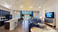 This single-floor clubhouse is designed to serve as a versatile gathering hub, combining comfort and functionality in a spacious, open layout. The entry foyer makes a strong first impression with soaring ceilings, transom windows, and French doors that flood the space with natural light, complemented by a decorative tile floor that transitions into warm hardwood throughout the interior.

The main social area centers around a large sectional sofa in bold navy blue, anchored by stylish area rugs and illuminated by a series of modern pendant lights suspended from the high ceiling. French doors and oversized windows along the exterior wall offer views of the surrounding greenery, enhancing the airy ambiance.

Adjacent to the lounge is a fully appointed kitchen featuring dark cabinetry, a tile backsplash, black appliances, and a generous white-topped island with bar seating — ideal for community events and gatherings. A water station nearby adds a practical amenity touch.

A separate lounge area provides a more intimate setting, furnished with rich leather club chairs arranged around a central bookcase display and ottoman, framed by dramatic floor-to-ceiling drapery panels. Flanking this space is a dedicated business center with individual computer workstations, offering residents a quiet and productive environment. The overall design blends approachable warmth with polished, contemporary style.