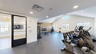 Located at 111 Sunset Circle in Camden, Delaware, this modern fitness center offers a thoughtfully designed single-floor layout dedicated entirely to health and wellness. The space is divided into two distinct areas: a main exercise room and an adjoining open studio, connected by striking black-framed glass French doors that lend a sleek, contemporary aesthetic throughout.

The main exercise room features soaring vaulted ceilings that amplify the sense of openness, complemented by large windows that invite abundant natural light and views of the surrounding area. The durable speckled rubber flooring provides both safety and style. Equipment is comprehensive and well-organized, including multiple treadmills, elliptical machines, a stationary bike, a cable strength training system, adjustable benches, a full dumbbell rack, medicine balls, and resistance bands. Wall-mounted flat-screen televisions add convenience for entertainment during workouts. A warm wood slat accent wall adds a refined, spa-like touch to the strength training zone.

The adjoining studio area features light hardwood flooring and a stunning gold tiered chandelier, creating an elegant yet functional space ideal for yoga, stretching, or guided fitness sessions. A large window brightens the room with natural light. Together, the two areas form a cohesive, upscale fitness facility that balances performance with sophisticated design.