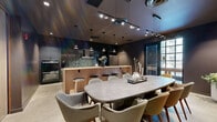 Located at 301 Winchester Ave in New Haven, CT, this sophisticated kitchen and dining space delivers a striking modern aesthetic defined by dark, moody tones and premium finishes throughout. The kitchen is outfitted with stainless steel appliances—including a large French-door refrigerator and built-in wall ovens—complemented by dramatic terrazzo countertops and a mosaic tile backsplash that adds texture and visual depth. A warm wood-toned island with leather-topped bar stools provides casual seating and serves as a natural dividing element between the kitchen and dining areas. A dedicated coffee bar station enhances the space's functionality. The dining area accommodates a generous oval concrete-top table surrounded by an eclectic mix of upholstered mid-century modern chairs in muted greens, grays, and taupes, creating an inviting gathering atmosphere. A wall of illuminated, glass-and-steel-framed display cabinetry anchors one side of the dining area, adding architectural elegance. Polished concrete flooring runs throughout, tying the design together cohesively. Layered lighting—including track lighting, recessed fixtures, and pendant lights—creates a dynamic and well-lit environment. Large windows allow natural light to filter in, offering views to the surrounding exterior.