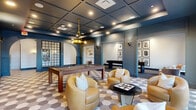 This thoughtfully designed single-floor clubhouse serves as a vibrant social hub, blending sophisticated aesthetics with recreational functionality. The main game area is anchored by a full-size billiards table beneath a striking brass pendant chandelier, complemented by a regulation shuffleboard table along the adjacent wall. Rich navy-blue walls, coffered ceilings with recessed lighting, and a distinctive geometric hexagonal tile floor establish a bold, upscale atmosphere throughout the tiled central zone.

Comfortable lounge seating in warm caramel leather is arranged throughout the space, encouraging casual conversation and relaxation. Built-in shelving with curated décor accents and framed black-and-white art gallery walls add a cultivated, editorial character to the interior. Arched doorways and decorative metal lattice partitions introduce architectural variety and visual depth.

Floor-to-ceiling windows along multiple walls flood the space with natural light while offering views of the surrounding landscaped grounds, outdoor amenity areas, and adjacent buildings. A quieter carpeted lounge nook with plush accent chairs provides a more intimate retreat within the larger space. The overall design balances energetic recreational programming with refined lounge comfort, creating a versatile and welcoming environment for socializing and leisure.