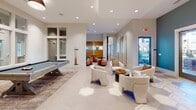 Located at 2818 Barrow Rd. in Charlotte, NC, this single-floor amenity space offers a thoughtfully designed environment built for both leisure and social interaction. The open-concept layout is anchored by large-format hexagonal tile flooring throughout, creating a cohesive and contemporary aesthetic across all areas.

The central gathering area features a full-size pool table set atop a layered area rug, surrounded by flexible seating arrangements that include round bistro tables paired with upholstered ottomans in warm blush and neutral tones. A natural wood accent wall with large mirror panels adds warmth and visual depth to the space.

Adjacent to the billiards area, a comfortable lounge zone provides curved upholstered armchairs and small side tables, bathed in natural light from multiple windows and glass-paneled doors that offer views of the surrounding outdoor landscape. Recessed lighting throughout ensures a bright, welcoming ambiance.

A dedicated kitchenette serves as a functional hub, equipped with a stainless steel beverage and ice machine, an under-counter refrigerator, a granite-topped counter with a sink, open wood shelving, and a white subway tile backsplash. Warm wood accents and recessed lighting complete the well-appointed space.

A separate break room, clearly marked by signage, offers a more intimate retreat with a glass partition window connecting it visually to the main lounge area.