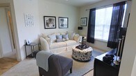 This single-floor residence offers a thoughtfully designed living space that blends comfort with contemporary style. The living room serves as the centerpiece of the home, showcasing a clean, neutral color palette of creams, grays, and blacks that creates a cohesive and sophisticated atmosphere. A plush cream sofa anchors the seating area, accessorized with an assortment of decorative pillows featuring geometric and floral patterns. A striking black-and-white patterned ottoman serves as both a coffee table and a focal point, complemented by a charcoal accent chair draped with a soft throw blanket. The space is finished with plush carpeting underfoot and a layered area rug that defines the seating arrangement. Natural light flows generously through large windows dressed with sleek black curtains and horizontal blinds, lending the room a bright and airy feel. Framed black-and-white artwork adorns the walls, adding a gallery-like touch to the overall aesthetic. Subtle accent lighting from a standing lamp and candleholders contributes to the warm, inviting ambiance. The overall design of this space reflects a modern sensibility with carefully curated décor, making it a comfortable and visually appealing environment.