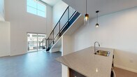 This contemporary two-floor residence makes a striking first impression with its dramatic double-height ceilings in the main living area, flooding the space with natural light through expansive floor-to-ceiling windows that frame urban views. Large-format tile flooring runs throughout the main level, giving the open-concept layout a clean, cohesive feel. A sleek staircase with a black metal railing ascends to the upper level, serving as both a functional element and a bold architectural focal point.

The kitchen is outfitted with dark wood cabinetry extending to the ceiling, stainless steel appliances including a full-size refrigerator and range, and a generous island featuring speckled quartz countertops with an integrated sink. Stylish pendant lighting hangs above the island, adding warmth to the modern aesthetic. A ceiling fan in the living area provides additional comfort.

The bathroom features a dark wood vanity with quartz countertops, an undermount sink, brushed nickel fixtures, and a large wall mirror. The upper level offers a carpeted loft-style space with a ceiling fan, open to the floor below via a railing, making it a versatile area well-suited for a bedroom or lounge. Additional carpeted rooms on this level include a bedroom with a walk-in closet, completing this well-appointed multi-level residence.
