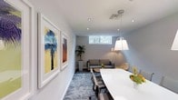 This thoughtfully designed single-floor space offers a refined and functional environment suited for professional use. The main area serves as a dual-purpose meeting and lounge space, anchored by a sleek white conference table paired with ergonomic task chairs, all set atop a patterned area rug that adds texture and visual warmth to the room.

A striking pendant light with a classic white shade hangs above the table, complemented by recessed ceiling lighting that ensures even, well-distributed illumination throughout. A floor lamp with a tri-arm design adds an additional layer of ambient light near the lounge corner.

Along one wall, two large framed art prints featuring bold tropical and abstract motifs introduce vibrant color and a contemporary aesthetic to the otherwise neutral palette of whites and soft grays. A tall potted plant in the corner brings a touch of nature indoors, enhancing the calm, welcoming atmosphere.

The lounge area at the far end features a gray sofa, an accent chair, and a small coffee table, creating an informal setting for conversation or relaxation. A horizontal clerestory window allows natural light to filter in, connecting the interior to the greenery outside. The overall ambiance is clean, modern, and professionally curated.
