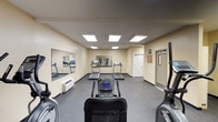 This fitness center is a dedicated single-floor exercise facility designed to accommodate a variety of cardio-focused workouts. The room features warm, neutral-toned walls paired with dark rubber flooring, creating a practical and comfortable environment for physical activity. Overhead, a series of recessed panel lights provide consistent, even illumination throughout the space.

The facility is outfitted with multiple treadmills, elliptical trainers, and a stationary recumbent bike, offering users a well-rounded selection of cardiovascular equipment. Large wall-mounted mirrors span portions of the room, visually expanding the space and allowing users to monitor their form during exercise. A wall-mounted HVAC unit ensures climate control year-round.

The layout is open and thoughtfully organized, allowing users to move freely between machines without feeling crowded. A standard door provides access to an adjacent room or corridor, suggesting convenient connectivity within a larger facility. Framed artwork on the walls adds a modest decorative touch to the otherwise functional space. Overall, this fitness center is clean, well-maintained, and efficiently designed to serve the exercise needs of its users.