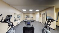 This fitness center is a dedicated single-floor exercise facility designed to accommodate a variety of cardio-focused workouts. The room features warm, neutral-toned walls paired with dark rubber flooring, creating a practical and comfortable environment for physical activity. Overhead, a series of recessed panel lights provide consistent, even illumination throughout the space.

The facility is outfitted with multiple treadmills, elliptical trainers, and a stationary recumbent bike, offering users a well-rounded selection of cardiovascular equipment. Large wall-mounted mirrors span portions of the room, visually expanding the space and allowing users to monitor their form during exercise. A wall-mounted HVAC unit ensures climate control year-round.

The layout is open and thoughtfully organized, allowing users to move freely between machines without feeling crowded. A standard door provides access to an adjacent room or corridor, suggesting convenient connectivity within a larger facility. Framed artwork on the walls adds a modest decorative touch to the otherwise functional space. Overall, this fitness center is clean, well-maintained, and efficiently designed to serve the exercise needs of its users.
