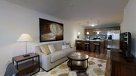 This thoughtfully designed single-floor space offers an open-concept layout that effortlessly blends the living, dining, and kitchen areas into one cohesive environment. The living area is anchored by a plush, neutral-toned sofa complemented by coordinating throw pillows and a round, metallic coffee table resting atop a patterned area rug in warm earth tones. A large framed artwork adds a bold focal point to the clean white walls, contributing to the space's curated aesthetic. Natural and ambient lighting work together to create a warm, inviting atmosphere throughout. The kitchen is outfitted with rich wood cabinetry, stainless steel appliances, and a teal accent wall that introduces a vibrant pop of color. Pendant lighting hangs above the kitchen bar, which is lined with sleek dark bar stools — ideal for casual dining or entertaining. The rich hardwood flooring flows throughout the open space, adding warmth and visual continuity. The overall design balances modern sophistication with comfortable, livable touches, making this a well-appointed space suited for both everyday living and hosting guests.