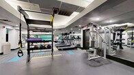 This expansive fitness center offers a comprehensive workout environment spread across a generously sized single floor. The space is thoughtfully organized into distinct training zones, each designed to support a wide range of exercise disciplines. The cardio area features a row of treadmills and elliptical machines, each equipped with individual media screens for an engaging workout experience. A dedicated strength training section provides a robust selection of selectorized weight machines, free weights, dumbbells, and adjustable benches, all arranged to allow ample space for movement. A functional training zone includes a multi-purpose rig with TRX suspension straps, medicine balls, foam rollers, and open floor space ideal for dynamic exercises and stretching. Flooring transitions between durable rubber and colorful patterned carpet tiles in teal, yellow, and grey, clearly delineating activity areas while adding visual interest. Floor-to-ceiling mirrors line multiple walls, enhancing the sense of space and allowing users to monitor their form throughout their workouts. Recessed and linear LED lighting illuminates the entire facility evenly. A tray ceiling detail in one section adds architectural dimension, while the overall design maintains a clean, modern aesthetic with teal accent walls providing a vibrant, energizing atmosphere throughout.