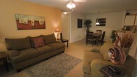 This single-floor residence offers an open and inviting layout that seamlessly connects the living and dining areas, creating a cohesive and functional living environment. The living area is anchored by plush, earth-toned seating arranged around a textured area rug, with a ceiling fan overhead providing both light and airflow. A warm accent wall in golden orange adds a pop of color and depth to the space, complemented by tasteful wall art. A closet door along the back wall suggests convenient built-in storage within the living area. Flowing naturally from the living space, the dining area accommodates a full dining table and chair set beneath a pendant light fixture, making it well-suited for everyday meals or entertaining. A decorative indoor plant and wall-mounted artwork add character to this zone. The consistent neutral carpet flooring throughout unifies the open floor plan, while the soft, layered lighting from ceiling fixtures and accent lamps creates a cozy and welcoming atmosphere. Overall, the space presents a practical and comfortable single-level layout with warm, residential finishes throughout.