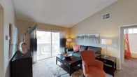 This single-story space centers around a well-appointed living room that combines comfort and functionality. The room features vaulted ceilings that lend an enhanced sense of openness, complemented by a textured ceiling finish and neutral warm-toned walls. A large sliding glass door serves as a prominent architectural feature, flooding the space with natural light while providing direct access to a private outdoor balcony, where outdoor seating can be seen. The balcony also offers views of surrounding greenery, adding a natural element to the interior atmosphere.

The living area is thoughtfully arranged with ample room for seating, anchored by a dark-finished wood coffee table with drawer storage. Neutral-toned carpeting with an area rug beneath the furniture grouping adds warmth to the floor. Dual-toned window treatments frame the sliding door, offering both light control and a design accent. An HVAC vent is visibly integrated into the wall, ensuring climate comfort. Overall, the space presents a bright, inviting atmosphere suitable for relaxed everyday living.