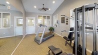 This single-story fitness facility is designed to provide a comfortable and functional workout environment. The main exercise room features vaulted ceilings with recessed lighting and a ceiling fan, contributing to a spacious, airy atmosphere. The walls are finished in a neutral gray tone, complemented by clean white trim and baseboards, while durable carpeted flooring runs throughout the space.

Large windows along multiple walls allow abundant natural light to fill the room, offering views of the surrounding outdoor area. A full-wall mirror spans one side of the exercise room, visually expanding the space and providing a practical training aid. Equipment includes a multi-station cable weight machine, a treadmill, an elliptical trainer, and a weight bench, offering a well-rounded range of cardio and strength training options. Instructional fitness posters are mounted near the equipment for user reference.

An interior pass-through window connects the fitness room to an adjacent area, maintaining visual openness between spaces. A restroom is conveniently accessible from within the facility, and a potted plant adds a touch of greenery to the overall setting.