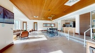 This thoughtfully designed single-floor space blends contemporary aesthetics with functional communal amenities. The entry area features a wood-and-white reception desk flanked by modern chairs, with large windows allowing abundant natural light and outdoor views. Warm wood-trimmed glass doors frame the entrance, establishing an inviting tone throughout.

The main open area is anchored by a striking wood-paneled tray ceiling with recessed lighting and a bold brass-and-globe pendant chandelier. Large-format tile flooring extends across the open layout, which is divided into distinct zones. A casual lounge area features leather seating arranged on a patterned area rug, while a raised dining zone centers on a high-top table with bar-height chairs. A step-up kitchenette in the background includes light wood cabinetry, dark tile backsplash, and appliances suitable for communal use.

A separate lounge room offers comfortable sectional and accent seating, large windows with exterior views, and curated artwork, creating a bright and relaxed gathering spot.

The carpeted corridor leads to a dedicated mail room lined with rows of individual mailboxes, complemented by a built-in wood service counter and coordinating patterned carpet, ensuring practicality alongside the space's refined design.