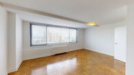 This single-floor unit presents a clean, minimalist design accented by warm parquet wood flooring that extends throughout the living and sleeping areas. The open layout, as revealed by the floor plan, organizes the space efficiently, with distinct zones for living, sleeping, kitchen, and bathroom functions.

The living area is notably bright, drawing in generous natural light through a wide bank of windows fitted with horizontal blinds. These windows frame elevated city views, offering a dynamic urban backdrop. A structural beam detail at the ceiling adds architectural character, while flush-mounted lighting fixtures provide consistent illumination.

The bedroom mirrors the living area's aesthetic, featuring the same parquet flooring and a window that captures additional natural light and outdoor views. A closet is accessible through a dedicated door, adding practical storage to the space.

The kitchen is compact yet functional, outfitted with stainless steel appliances including a gas range and an over-the-range microwave by Frigidaire. Light wood cabinetry pairs with crisp white countertops and an undermount stainless steel sink with a modern arc faucet. An electrical panel is conveniently located nearby.

The bathroom maintains a simple, clean aesthetic with white tile surrounds, a full bathtub, and straightforward fixtures throughout.