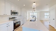 This thoughtfully designed three-floor home offers a clean, modern aesthetic defined by crisp white walls, light hardwood flooring, and an abundance of natural light throughout. The ground level houses a spacious garage with concrete flooring, interior access, and an electrical panel, providing both storage and convenience. Carpeted staircases with warm wood handrails connect the floors seamlessly.

The main living floor features an open-concept kitchen and living room, where white shaker cabinetry, quartz countertops, and stainless steel appliances — including a range, microwave, and refrigerator — surround a central island with an undermount sink and a striking matte black faucet. Pendant lighting accents the island, while large windows at the end of the space flood the living area with natural light and neighborhood views. Light hardwood floors extend throughout, enhancing the bright, airy atmosphere.

The upper floor is home to three bedrooms, each featuring hardwood flooring, recessed lighting, ceiling fans, and windows offering outdoor views. The primary bedroom includes an attached bathroom with white cabinetry, a single-sink vanity, and a dedicated closet with built-in wire shelving. The additional bathrooms mirror the same clean, contemporary design, maintaining a cohesive style across the entire home.