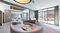 Located at 2870 Pearson Pkwy in Plainfield, Indiana, this thoughtfully designed single-floor amenity space offers a compelling blend of collaborative and private environments across a generous footprint. The layout comprises multiple lounge areas, two conference rooms, private office nooks, and a dining area — each distinct in character yet unified by a cohesive contemporary aesthetic.

The main lounge welcomes visitors with curved, modular seating arrangements atop vibrant mixed-tone carpeting, complemented by exposed wood beam ceiling details and statement pendant lighting. Floor-to-ceiling windows flood the space with natural light and provide views of the surrounding community. A high-top bar counter with leather stools lines the windowed perimeter, creating an inviting casual seating zone.

The conference rooms are well-appointed, each featuring a substantial wood-topped table, ergonomic seating, and bold globe pendant fixtures. One room showcases built-in shelving with curated décor and a private booth nook, while the other offers interior glass windows that maintain visual connectivity. A compact private workstation with a marble-surfaced desk and designer wall sconce provides a focused solo workspace.

The dining area features dark cabinetry with brass hardware set against a richly textured tile accent wall. Throughout, the space is accented with original artwork, lush greenery, and a carefully curated palette of gray, warm wood, and rich amber tones.