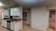 This single-floor space offers a practical, open-concept layout that connects the kitchen and living areas seamlessly. The kitchen features white cabinetry with clean-lined cabinet doors, a dark countertop that provides a subtle contrast against the lighter elements, and a peninsula-style pass-through that visually links the kitchen to the adjoining living space. This design choice encourages an open flow between the two areas while maintaining a defined cooking zone.

The living area is finished with wall-to-wall carpet in a warm, neutral tone, contributing to a comfortable and understated ambiance. The walls throughout are painted in a soft beige, creating a cohesive, calming palette that complements the flooring. A wall-mounted heating vent is positioned at the base of the wall, providing efficient climate control. Recessed lighting in the living area adds functional illumination, while a window at the far end of the space allows natural light to filter in, offering a view of outdoor greenery. The overall design is straightforward and functional, with a neutral aesthetic that provides a versatile backdrop for a variety of interior styles.