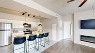 This single-story unit in Tulsa, OK offers a thoughtfully designed layout across a compact yet functional footprint. The open-concept living and kitchen area serves as the heart of the space, where wood-look flooring, neutral wall tones, and large windows create a bright, airy atmosphere. The kitchen is outfitted with high-gloss white cabinetry extending to the ceiling, stainless steel appliances, a tile floor, and a peninsula with counter seating highlighted by stylish black-and-gold pendant lighting. The adjoining living room features wood-look flooring, a built-in electric fireplace, a ceiling fan, and large windows that invite natural light and outdoor views. The spacious primary bedroom is finished with plush carpeting, a ceiling fan, natural light from dual windows, and direct access to a walk-in closet. The primary bathroom offers a wide double-sink vanity with backlit mirrors, crisp white cabinetry, and a combined tub and shower with tile surround. A dedicated laundry area with tile flooring and built-in shelving adds practical convenience to the overall layout.