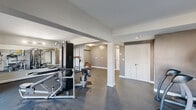 Located at 879 West Blvd in Hartford, Connecticut, this fitness center offers a thoughtfully designed exercise space across a single floor. The room features a durable dark-toned flooring throughout, providing a practical and resilient surface well-suited for a range of workout activities. Neutral gray walls and white trim give the space a clean, modern aesthetic, while structural columns divide the area into distinct workout zones without sacrificing an open feel. Large floor-to-ceiling windows along one side of the room flood the space with natural light and provide views of the surrounding neighborhood, creating an energizing atmosphere. Full-length wall mirrors in one section enhance the sense of depth and allow users to monitor their form during exercise. The facility is equipped with a multi-station cable weight machine, a bench press, an elliptical trainer, a stair climber, a treadmill, and a flat-screen television mounted on the wall for entertainment during workouts. A dedicated storage closet and additional utility areas are accessible via white double doors. Overhead flush-mount lighting ensures the space remains well-lit throughout. The overall layout is functional and efficient, offering a well-rounded environment for both cardiovascular and strength training.