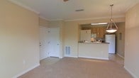 This single-floor space showcases an open-concept design that seamlessly connects the living area to the kitchen. The living area features neutral beige carpeting and warm-toned walls, complemented by crown molding that adds a refined architectural detail along the ceiling perimeter. A ceiling fan is centrally positioned, contributing to both comfort and airflow.

The kitchen is accessible via a raised breakfast bar counter, which serves as both a functional workspace and a visual divider between the two areas. Light wood cabinetry lines the kitchen walls, paired with under-cabinet lighting that illuminates the countertops below. A full-size refrigerator and microwave are visible among the appliances, and pendant lighting adds a decorative touch above the kitchen space.

Two white panel doors are visible near the entryway, likely providing access to closets or utility areas. A floor-level air vent is built into the baseboard area, indicating a central HVAC system. White baseboards frame the room cleanly, reinforcing the overall cohesive and well-maintained appearance of the space.