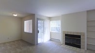 This single-story space features a clean, functional layout centered around a well-lit living area. The room is finished with neutral wall tones and light-colored carpeting, creating a calm and understated atmosphere throughout. A standout feature is the tile-surround fireplace positioned along one wall, adding a classic focal point to the living space. The fireplace features a dark insert framed by beige ceramic tile, complementing the room's neutral palette.

Natural light is a defining characteristic of the living area, which benefits from multiple windows fitted with horizontal blinds, as well as a sliding glass door dressed with vertical blinds. The sliding door provides direct access to an outdoor area and contributes significantly to the brightness of the interior. A baseboard heating unit is visible beneath one of the windows, indicating a reliable in-unit heating system. The ceiling is smooth and white, reinforcing the open and airy feel of the space. Overall, the living area presents a straightforward, move-in-ready environment with practical features and ample natural light.