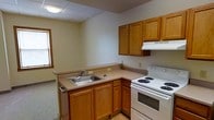 This single-floor space centers around a functional kitchen designed for efficient use of space. The kitchen features warm-toned wood cabinetry, including upper wall-mounted cabinets and lower base cabinets that provide ample storage. The countertops follow an L-shaped configuration, offering practical workspace for meal preparation. A stainless steel single-basin sink is integrated into the countertop along one wall, accompanied by a standard faucet. A white electric coil range with an oven occupies the adjacent wall, topped by a built-in vent hood with an upper cabinet above. The ceiling is finished with a suspended drop-tile grid system and includes a flush-mounted light fixture that provides overhead illumination. A small window along one wall allows for natural light to enter the space. The flooring transitions from a textured surface near the kitchen base to a lighter material near the range area. The neutral wall color and clean layout give the kitchen a straightforward, utilitarian character suited to everyday use.