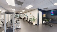Located at 14175 Castle Blvd in Silver Spring, Maryland, this fitness center offers a well-appointed, single-floor exercise space designed to support a full range of workout routines. The room features a drop-panel ceiling with recessed fluorescent lighting that keeps the space evenly and brightly illuminated throughout. Durable rubber flooring in a dark speckled finish covers the entire area, providing both safety and comfort underfoot.

The cardio zone is highlighted by large floor-to-ceiling windows that invite natural light and offer views of lush greenery outside, creating an energizing workout environment. Treadmills, elliptical trainers, and a recumbent bike are arranged to take full advantage of this natural light. Full-length mirrors are positioned along key walls, visually expanding the space and allowing users to monitor their form.

The strength training section includes a cable machine, a multi-function pulley system, a flat bench, and a rack of free weights with dumbbells and kettlebells neatly organized on tiered racks. A dedicated spin bike area features a wall-mounted smart TV and dark accent walls, lending the corner a boutique fitness studio feel. Colorful artwork adds a welcoming, motivating touch to the neutral white and charcoal color palette. A wall-mounted television and a connected restroom further enhance the functionality and convenience of this comprehensive fitness facility.