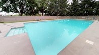 The outdoor pool area offers a generous, rectangular swimming pool filled with clear, turquoise water, creating an inviting and refreshing atmosphere. The surrounding concrete deck provides ample space for relaxation and movement, lined with several lounge chairs positioned to catch sunlight throughout the day. A privacy fence encloses the entire area, offering seclusion while mature trees rise beyond the perimeter, adding natural shade and a lush green backdrop. The combination of the well-maintained pool, open deck space, and verdant surroundings creates a calm, resort-like setting ideal for leisure and recreation.