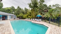 Located at Bristol Lakes in Mount Dora, Florida, this single-story community amenity facility offers a well-appointed outdoor retreat designed for relaxation and recreation. The centerpiece of the space is a generously sized swimming pool with clear turquoise water, surrounded by a broad paver brick deck that provides ample room for lounging. Multiple white sling-style chaise lounges are arranged along the poolside, complemented by a vibrant blue umbrella for shade, making it an ideal setting for sun-soaked afternoons.

The clubhouse building itself is architecturally distinctive, featuring a prominent cupola with a clock tower, a steeply pitched roof, and a welcoming covered entrance with clean white columns and French doors. The exterior blends neutral stucco with blue lap siding accents, creating a polished yet approachable aesthetic. A covered walkway with recessed lighting runs along the pool-facing side, offering a shaded transition between the building and the pool deck.

The entire pool area is enclosed by a black iron fence, providing both security and an unobstructed view of the lush, mature Florida landscape beyond — with tall palm trees, oaks, and dense greenery creating a natural, private backdrop that enhances the overall ambiance of the outdoor space.