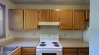 Spanning two floors, this space offers a practical and straightforward layout. The kitchen is equipped with warm oak wood cabinetry that lines the upper walls, providing generous storage space. A white electric coil-burner range serves as the focal point of the cooking area, complemented by a ventilation hood mounted beneath the upper cabinets. The lower cabinetry matches the upper units in finish, creating a cohesive look throughout the kitchen. The walls are painted in a neutral light gray tone, contributing to a clean and understated aesthetic. A textured ceiling adds a subtle visual element overhead. The kitchen's design prioritizes functionality, with ample counter and cabinet space for everyday use. The overall ambiance of the space is practical and comfortable, with classic finishes that offer a timeless, no-frills appeal. The two-floor layout suggests additional living, sleeping, or utility areas beyond the kitchen, providing versatile space for a variety of uses.