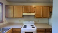 Spanning two floors, this space offers a practical and straightforward layout. The kitchen is equipped with warm oak wood cabinetry that lines the upper walls, providing generous storage space. A white electric coil-burner range serves as the focal point of the cooking area, complemented by a ventilation hood mounted beneath the upper cabinets. The lower cabinetry matches the upper units in finish, creating a cohesive look throughout the kitchen. The walls are painted in a neutral light gray tone, contributing to a clean and understated aesthetic. A textured ceiling adds a subtle visual element overhead. The kitchen's design prioritizes functionality, with ample counter and cabinet space for everyday use. The overall ambiance of the space is practical and comfortable, with classic finishes that offer a timeless, no-frills appeal. The two-floor layout suggests additional living, sleeping, or utility areas beyond the kitchen, providing versatile space for a variety of uses.