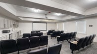 This screening and presentation room offers a thoughtfully designed, single-floor layout that comfortably accommodates an audience in a theater-style arrangement. The space features rows of cushioned, armrest-equipped seats with built-in cup holders, oriented toward a large projection screen mounted at the front of the room. A ceiling-mounted projector provides the AV capability, making the space well-suited for presentations, film screenings, or group gatherings. Crown molding and a coffered ceiling detail add architectural refinement to the otherwise clean, neutral interior, with white walls and patterned carpet contributing to a professional yet comfortable ambiance. Along one wall, a full built-in kitchenette includes gray shaker-style cabinetry, a sink, microwave, and under-counter refrigerator, offering convenient refreshment access. Additional flexible seating — including upholstered accent chairs in both navy blue and neutral tones — lines the perimeter, allowing for varied configurations. Multiple windows fitted with roller shades provide controlled natural light, while flush-mounted ceiling fixtures ensure consistent illumination throughout. The room strikes a balance between functional utility and polished design, making it a versatile venue for community screenings, lectures, or private events.